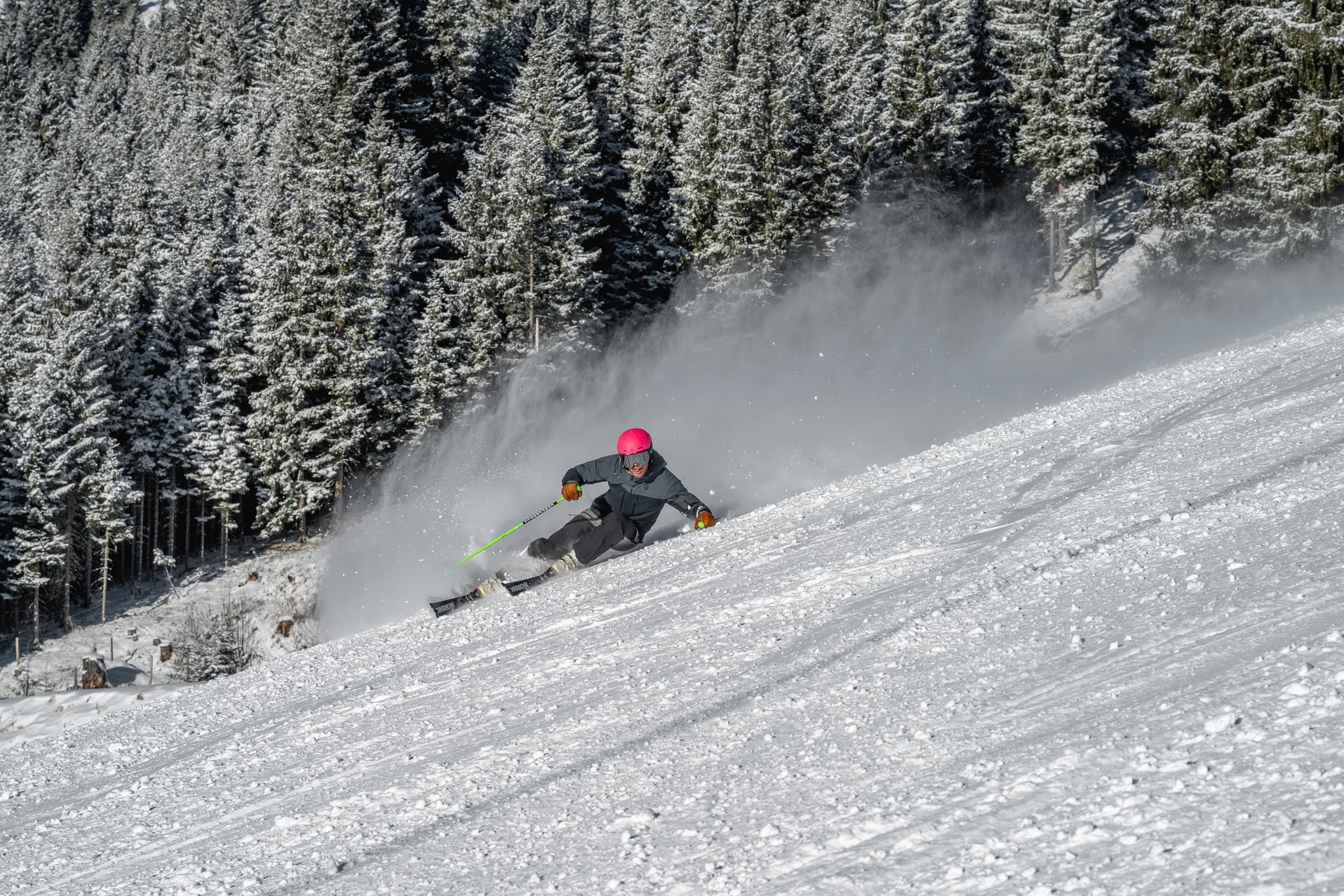 A skier carving a fast turn on a groomed slope with snow covered pines behind