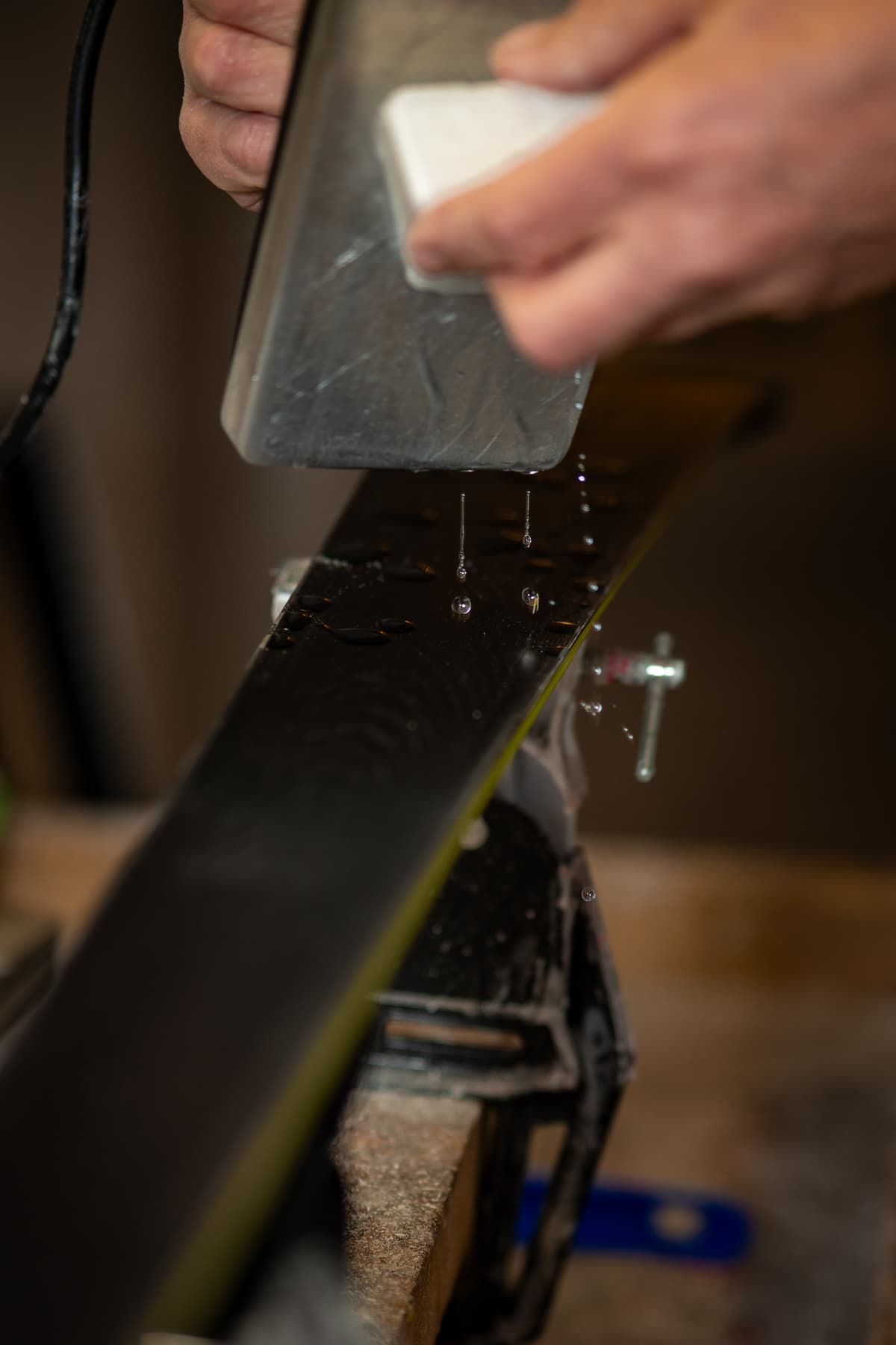Hands drawing a metal scraper along a freshly waxed ski base in a workshop