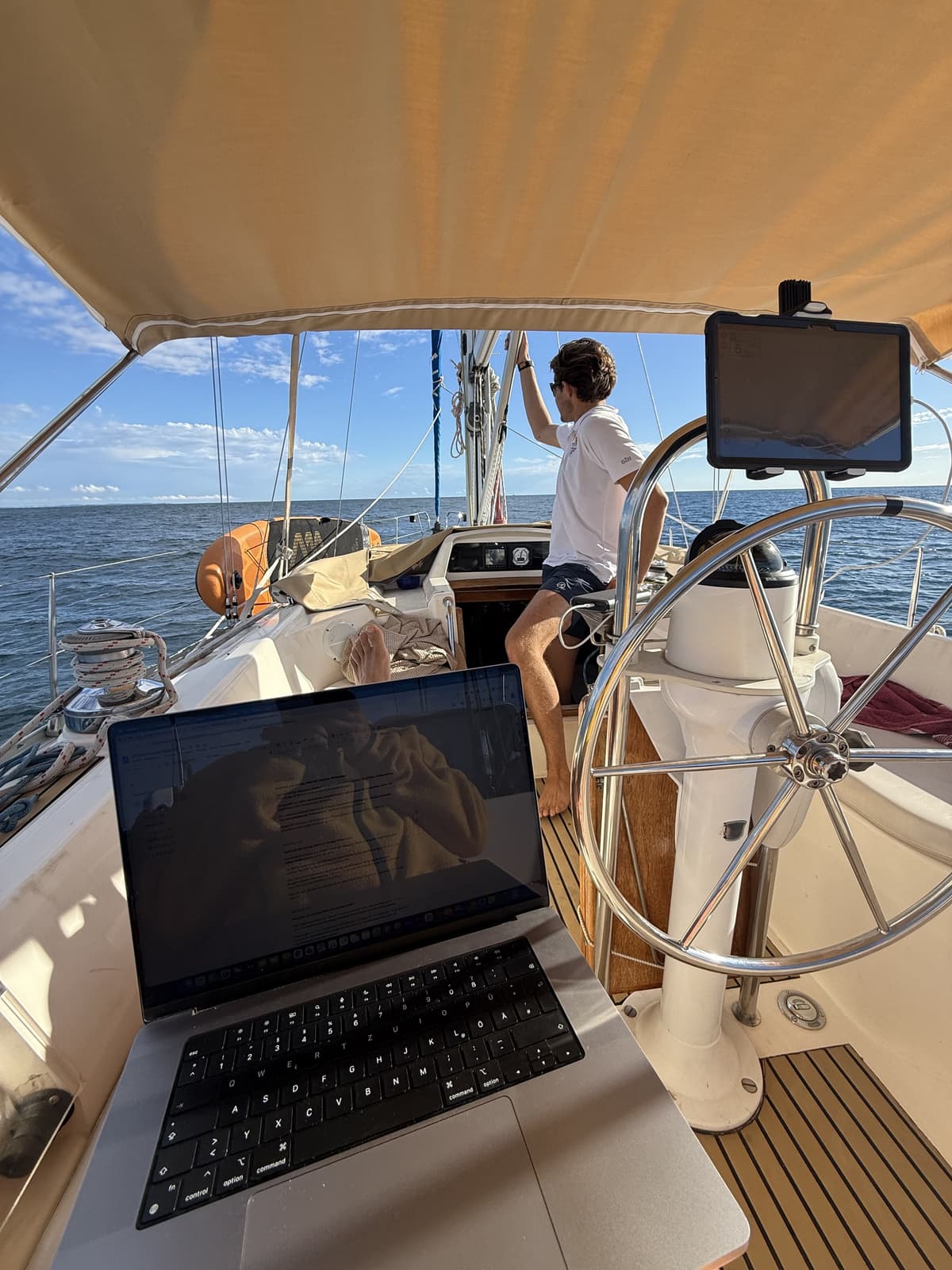 An open laptop on the cockpit table of a sailing yacht under way, with the helmsman standing at the wheel