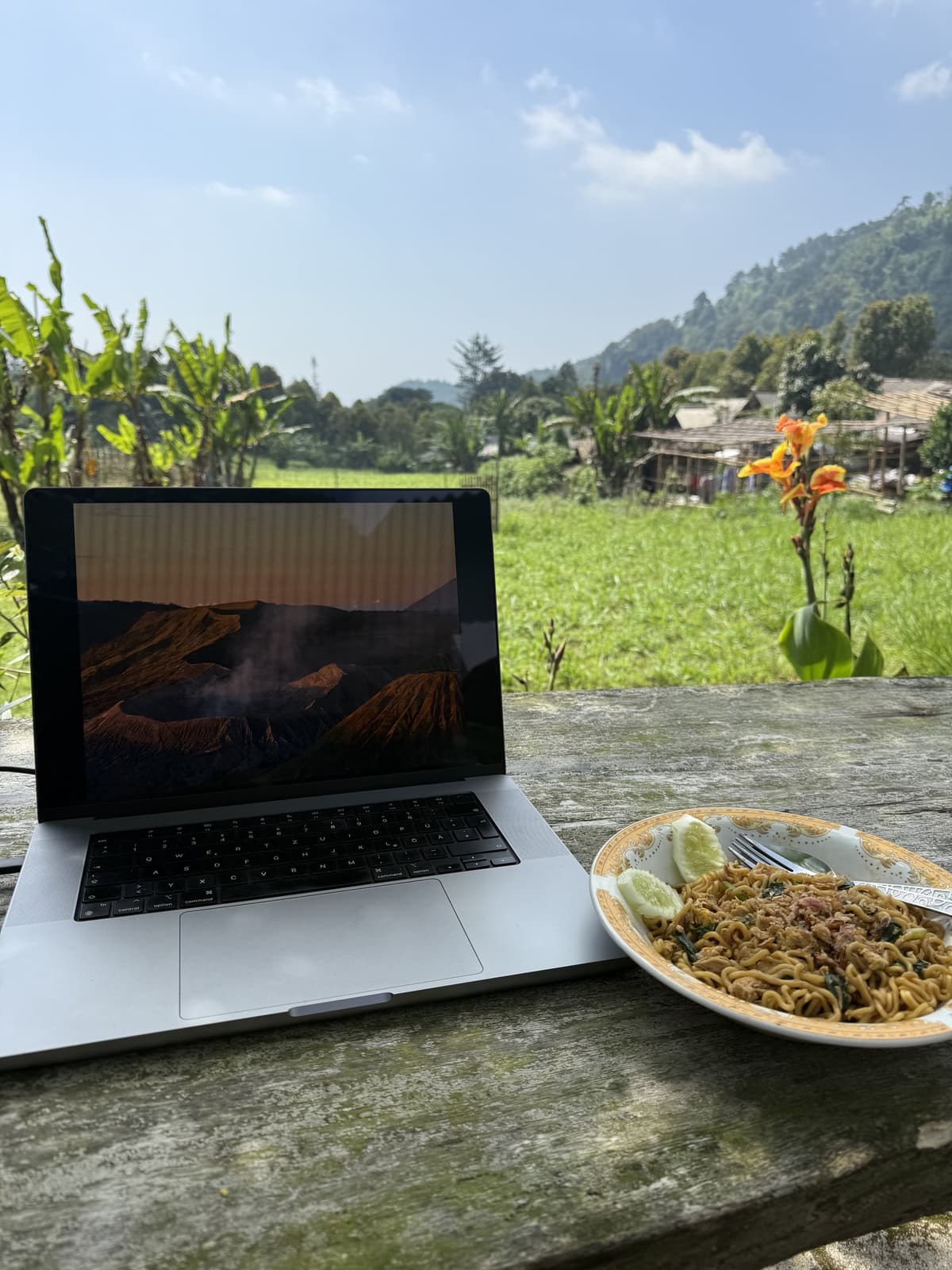 A laptop and a plate of noodles on a weathered wooden table looking out over green fields and hills in Indonesia