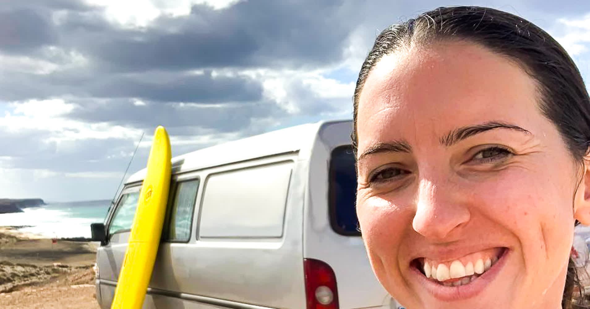 Lara Lain, remote marketer and digital nomad, smiling beside her surf van and yellow surfboard on a Fuerteventura beach during a workation.
