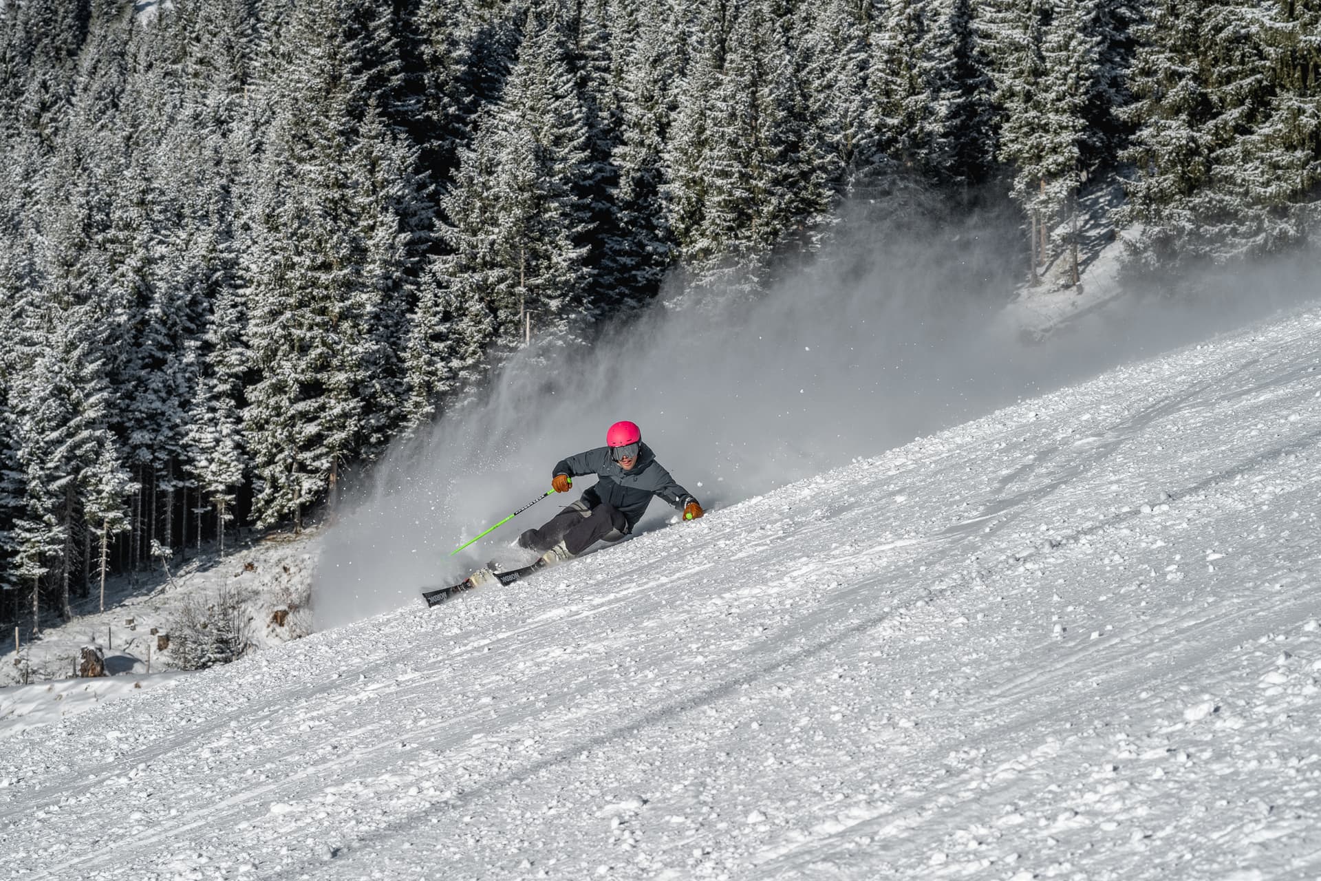 A skier in a red helmet carving a turn on a groomed piste, throwing up a plume of snow, with snow covered conifers on the slope behind.