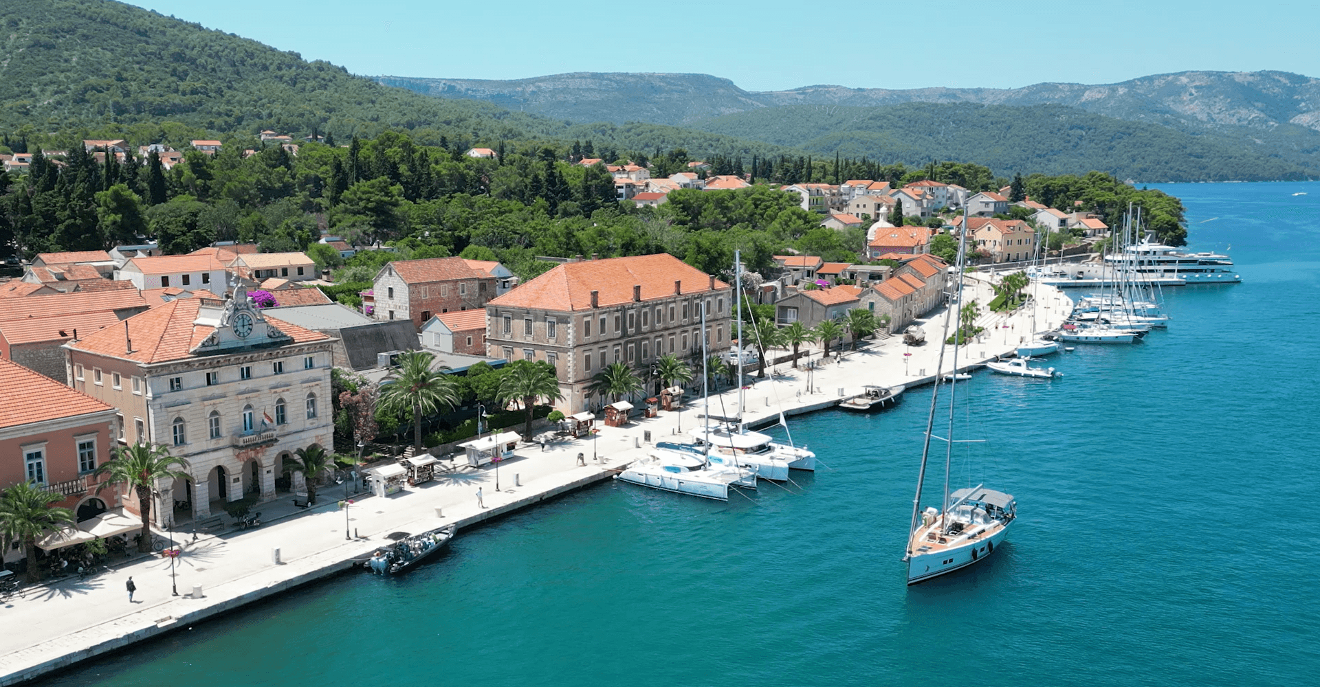 Aerial view of a Croatian coastal town harbour, sailing boats moored along a palm-lined stone promenade